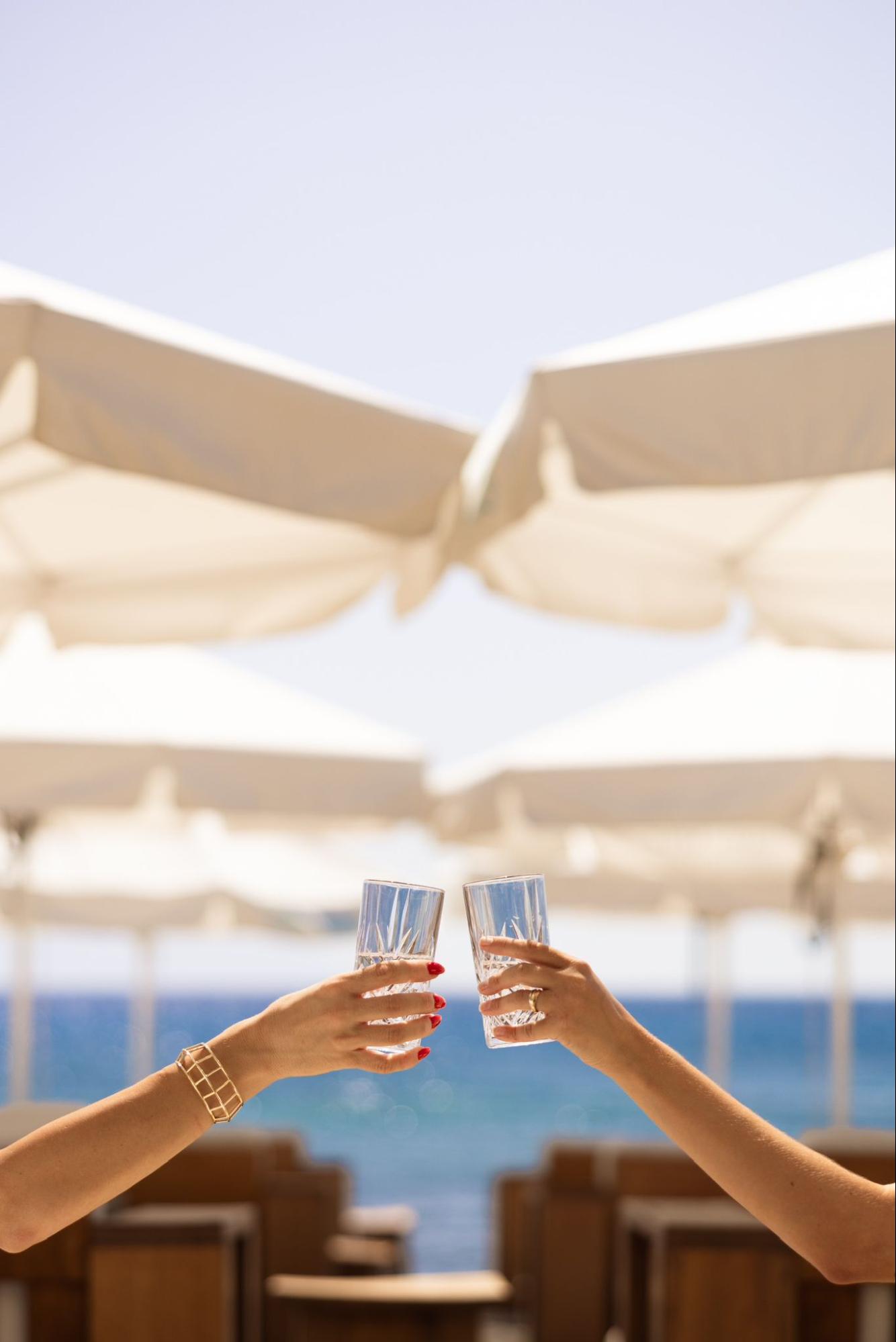 Two hands clinking glasses in a sunny toast beneath white beach umbrellas, with the bright blue ocean in the background, perfectly capturing the feel-good energy and connection of the signature Amazing Sundays experience