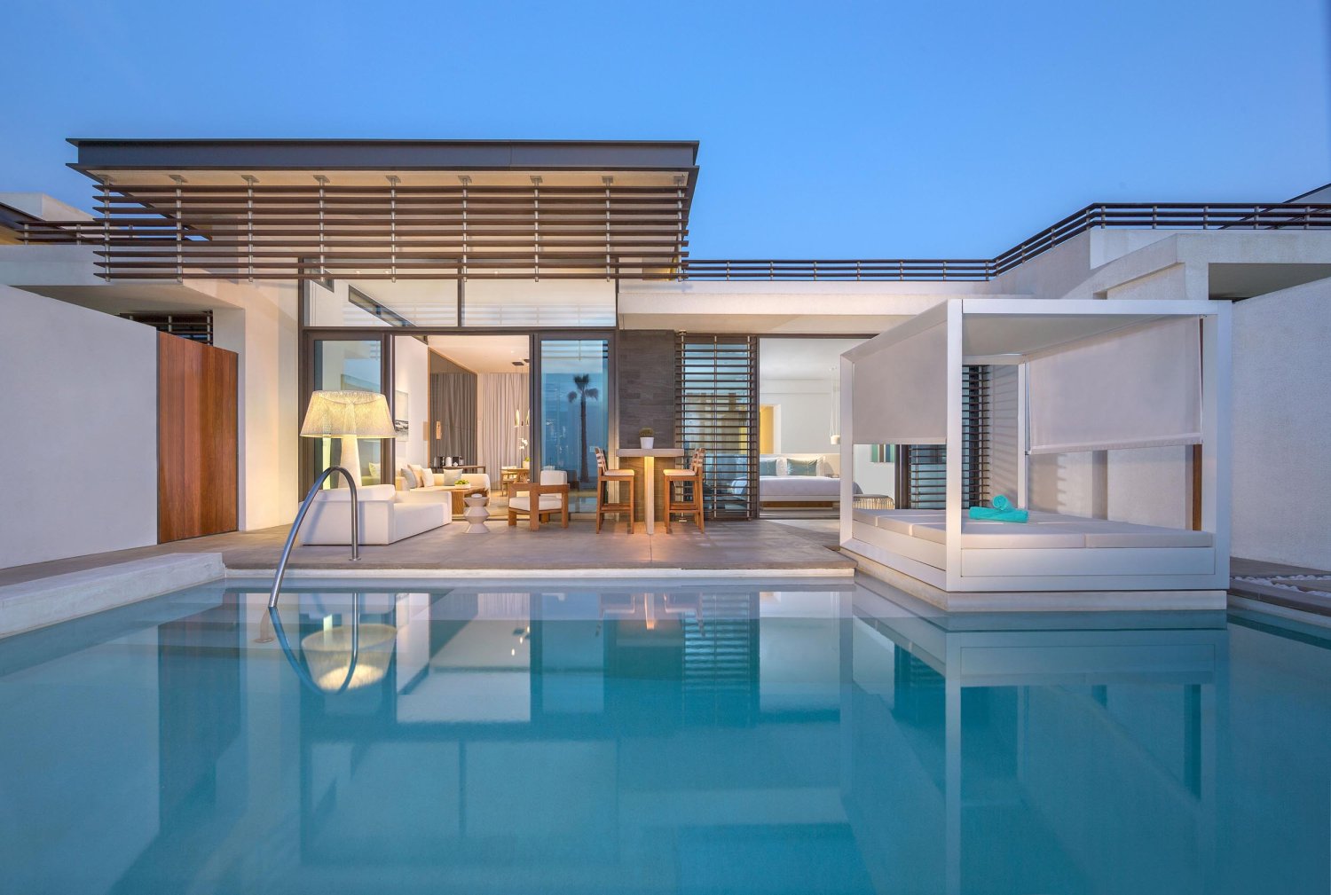 A view across the pool of the Beach villa, showing the sun bed, outside seating areas. Sliding doors are open, showing the living room areas inside