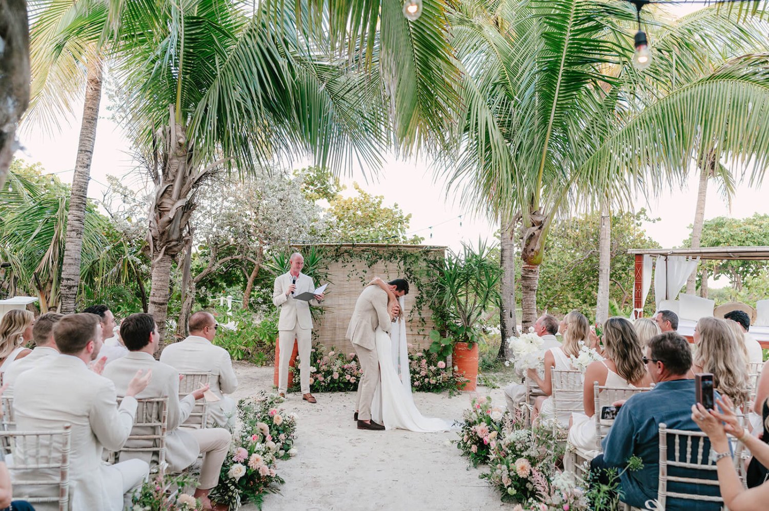 A couple hugging after getting married on a beach, surrounded by family with palm trees in the background,