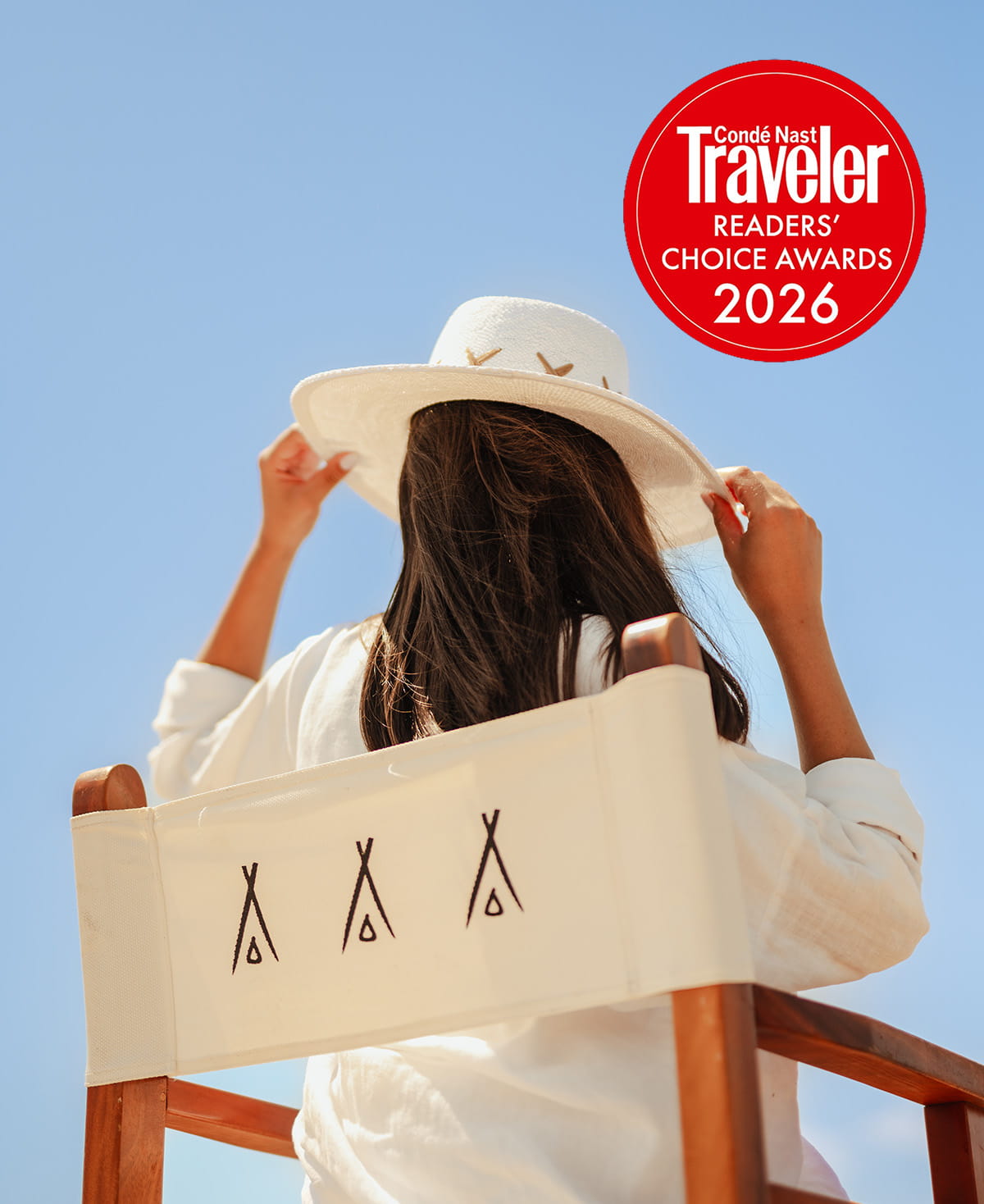 A woman seen from behind, sitting in a wooden director's chair featuring the signature Nikki Beach teepee logo. She is holding the brim of her white sun hat against a clear, bright blue sky, with a red Condé Nast Traveler Readers' Choice Awards 2026 badge prominently displayed in the top right corner to accompany the message asking for guest votes