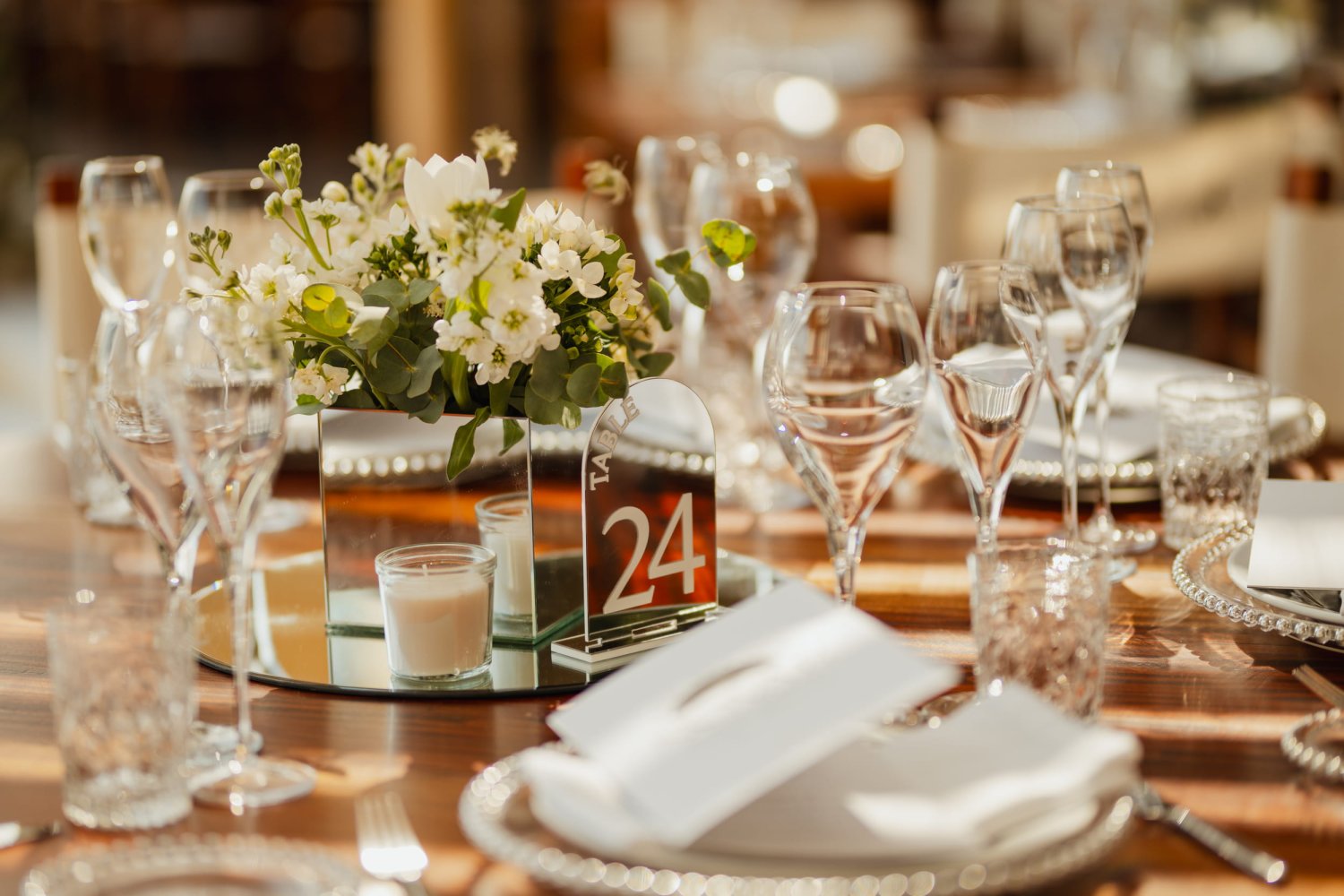 A beautifully arranged dining table bathed in warm sunlight, set for a private event or sophisticated reception. The polished wooden table features silver beaded charger plates, elegant glassware, and a mirrored centerpiece holding a fresh arrangement of white flowers and eucalyptus. An acrylic table number 