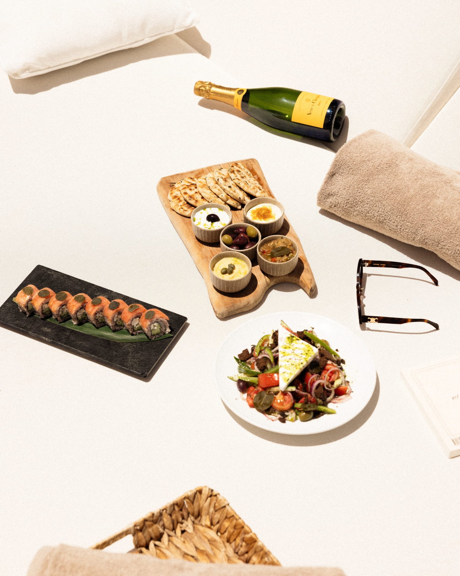 A luxurious dining spread laid out on a plush white daybed, featuring a fresh Greek salad, a wooden mezze board with warm pita bread and assorted dips, a plate of savory sushi rolls, and a chilled bottle of champagne. This elegant presentation captures the vibrant coastal cuisine and lively atmosphere of Nikki Beach Santorini
