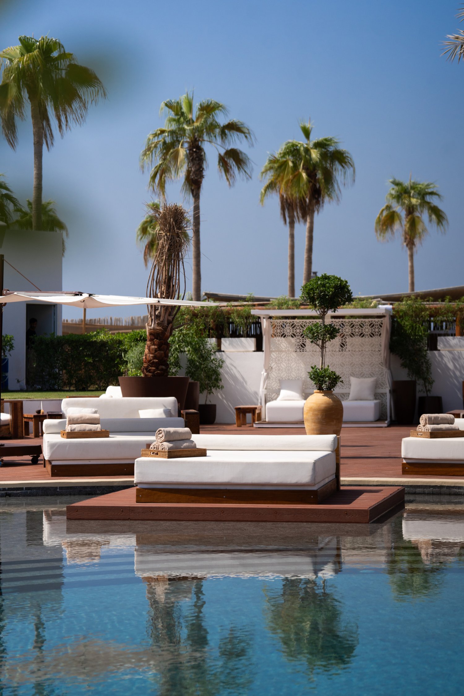 A serene view of the pool area at Nikki Beach Dubai, featuring a pristine swimming pool that beautifully reflects the surrounding tall palm trees and clear blue sky. Luxurious white daybeds complete with rolled towels rest on wooden decking, including one perfectly positioned over the water, capturing the sophisticated and relaxing ambiance of this Pearl Jumeira hotspot